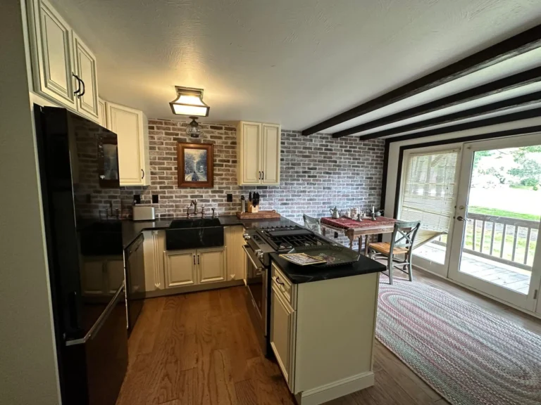 renovated kitchen with cream cabinets, black countertops, brick backsplash, exposed ceiling beams and a central cooking peninsula